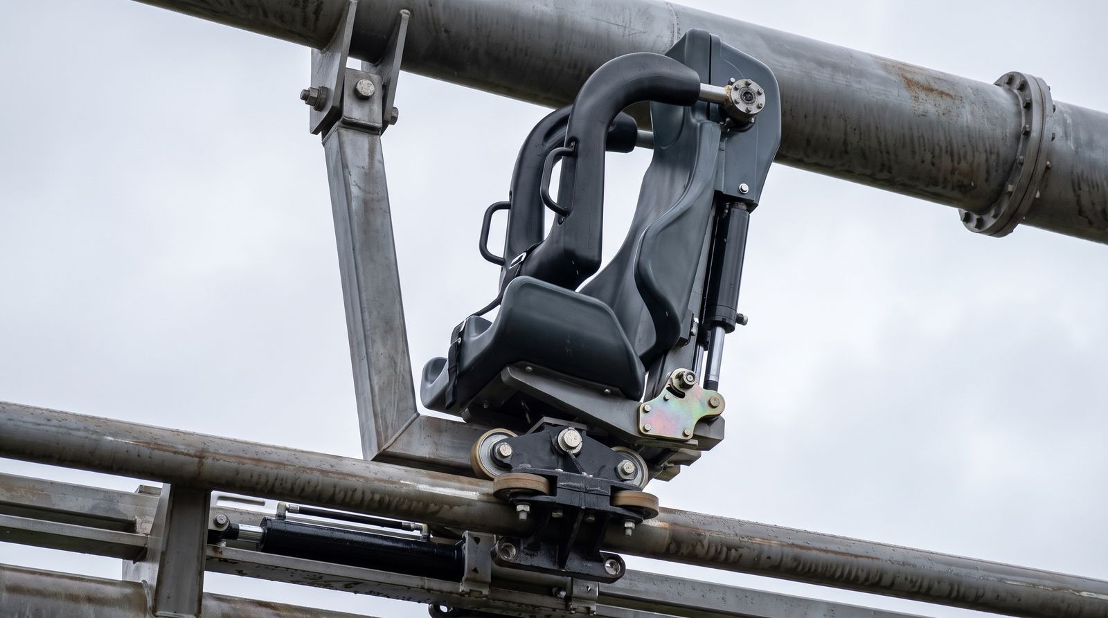 Close-up of a Bolliger and Mabillard wing coaster seat pod cantilevered to the side of a tubular steel rail, showing the wheel truck beneath the mounting bracket and a vest-style restraint with side grab handles
