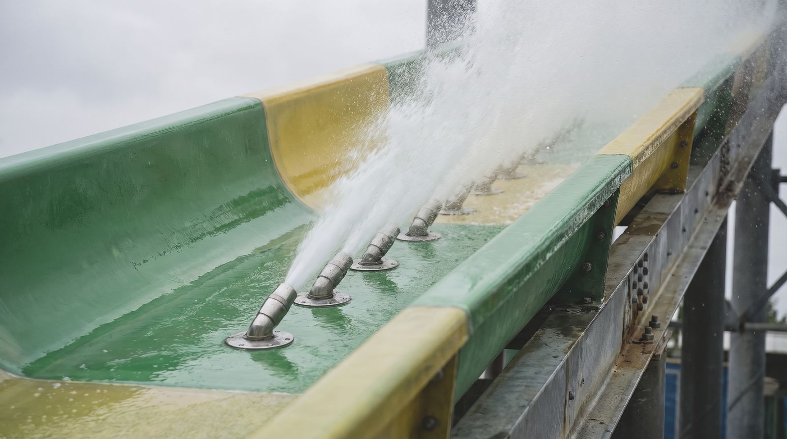 Close-up of a row of angled steel water jet nozzles in the floor of a green and yellow fibreglass flume, firing a powerful spray up the channel