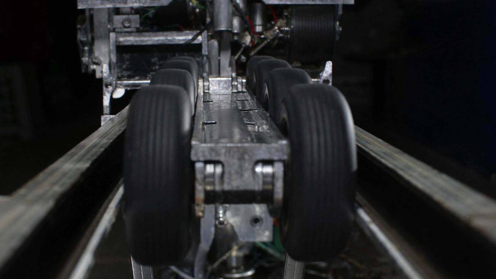 Close-up along a roller coaster booster drive section: paired black rubber tyres in steel frames gripping a central spine between the running rails
