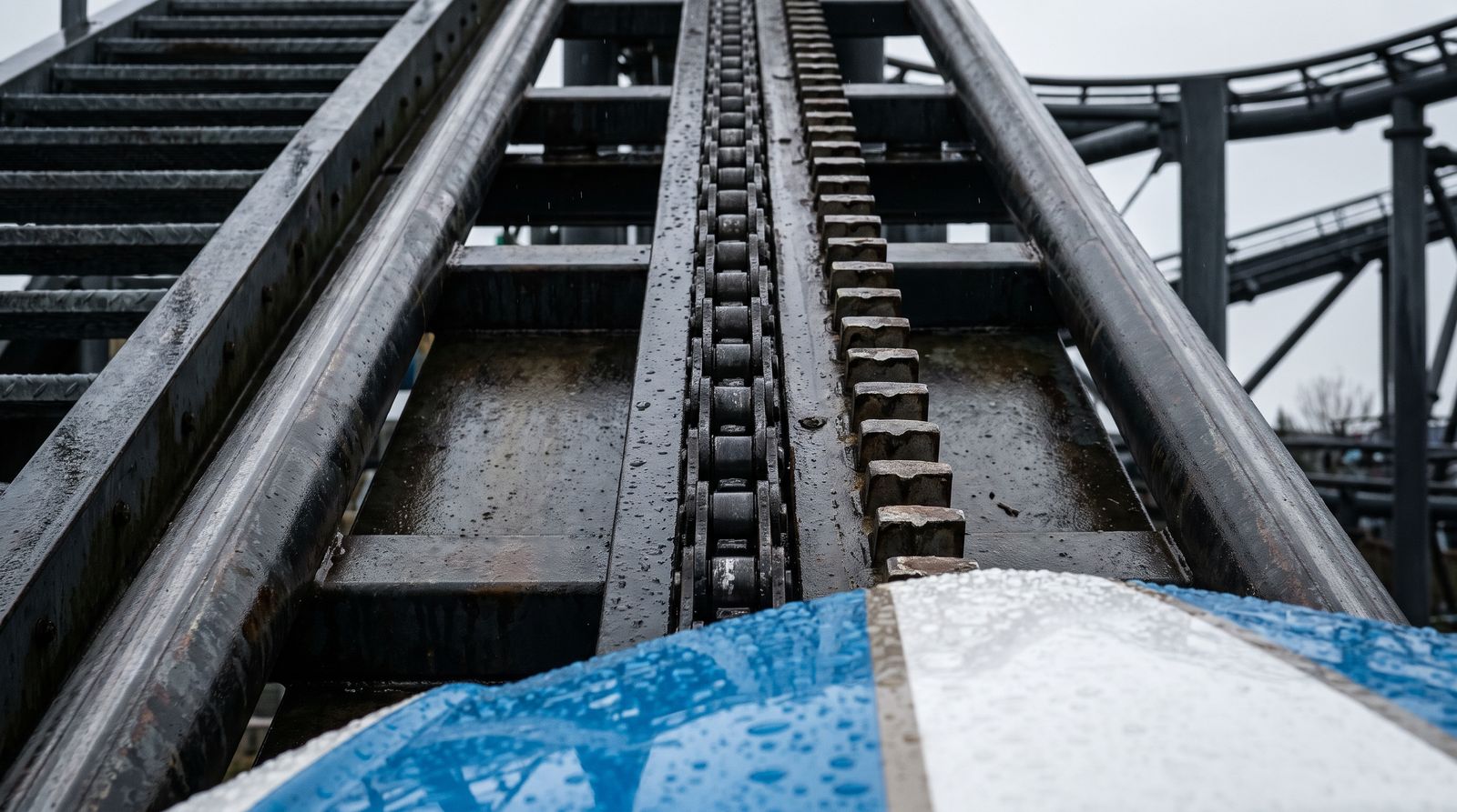 Close-up of a wet chain lift hill: a heavy link chain between the rails, a toothed anti-rollback rack beside it and the bow of a blue and white boat at the bottom of the frame